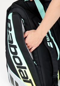 Hånd lægger en genstand i den sidelukkede lomme på en sort og neon Babolat sportstaske med skulderremme.