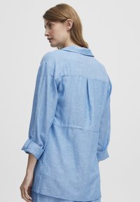 Gaišzils lina krekls ar aizmugures jostu, sarullētām piedurknēm un apkakli. Tam ir brīvs piegriezums un smalka tekstūra.