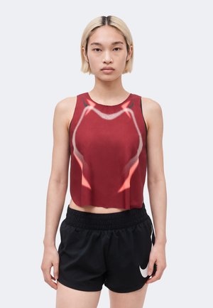 Person med kort blondt hår iført en rød ærmeløs crop top og sorte sportsshorts med hvidt Nike-logo, vendt fremad.