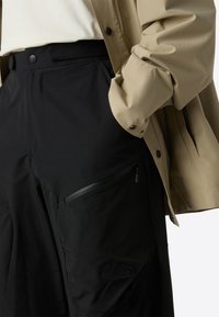 Crne cargo hlače s džepom na zip, u kombinaciji s bež vodootpornom jaknom, koja ima mat finiš i kopčanje na gumbi.
