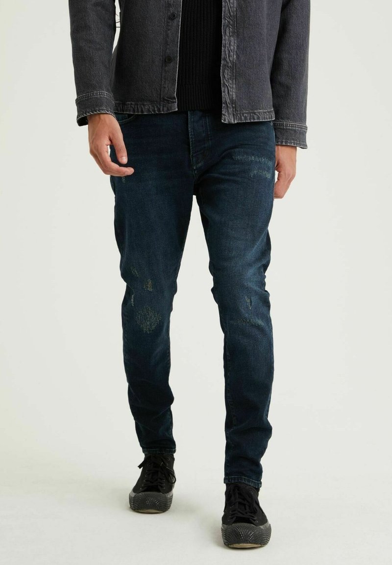 Mörkblå denimjeans med slitna lappar, slim fit och subtila slitningar. Kombineras med svarta sneakers och en mörk knopplös skjorta.