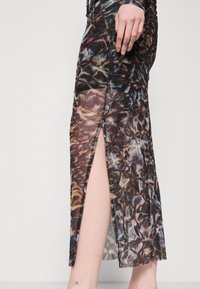 Femme portant une robe foncée à motifs avec une fente latérale dévoilant sa jambe, des tatouages visibles sur les doigts et la cheville.