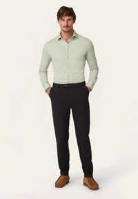Lichtgroen langslavend shirt, zwarte broek, bruin suède instap schoenen. Het shirt heeft knopen en heeft een getailleerd ontwerp.