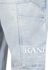 Gaiši zila denima kravas bikses ar lielu sānu kabatu un balto izšūto "KANI" logo zem tā. Mīksta tekstūra, standarta piegriezums.