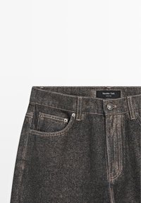 Jean en denim noir avec taille haute, comportant cinq poches, des coutures beige et une fermeture à bouton en métal. Étiquette de la marque visible au niveau de la taille.