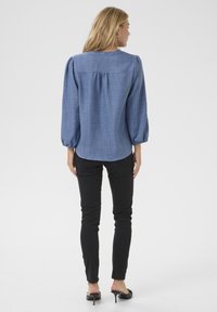 Blusa in chambray blu con maniche a sbuffo, dettaglio arricciato sulla schiena e scollatura rotonda, abbinata a pantaloni neri slim-fit e sandali con tacco.
