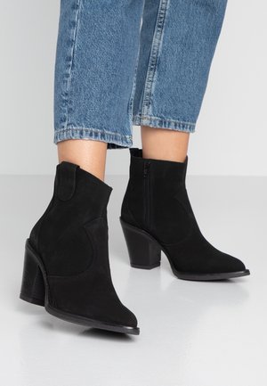 Person iført blå cowboybukser i midterlængde og sorte ruskindede ankelstøvler med blokhæle, stående på en glat, lysefarvet gulv.