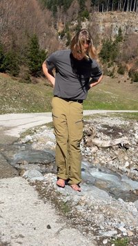 Personne en chemise grise et pantalon beige marchant pieds nus dans un ruisseau de montagne peu profond avec des berges rocheuses et un versant boisé en arrière-plan.