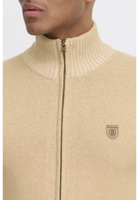 Suéter beige con cremallera y cuello alto acanalado, que presenta una textura sutil y un parche de logo en el lado izquierdo. La cremallera en la parte delantera añade un toque de detalle.