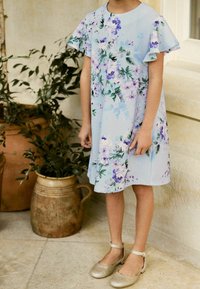 Helles blaues Kleid mit floralem Muster in Lila und Grün. Kurze Rüschensäume. Ein Paar goldene Schuhe mit Knöchelriemen.