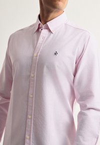 Camisa de botón de color rosa claro con finas rayas blancas, cuello puntiagudo y un logo bordado en el lado izquierdo del pecho. Los botones son de color crema.