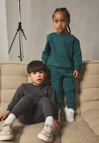 Deux enfants sur un canapé beige texturé. L'un porte un sweat-shirt et un pantalon turquoise ; l'autre porte une tenue gris foncé avec des baskets blanches.
