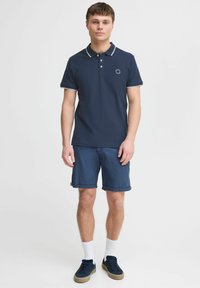 Námornícky polo s bielym lemovaním, krátkymi rukávmi a trojitém gombíkovom zapínaní, skombinovaný s zvitými džínovými šortkami a tmavými teniskami.