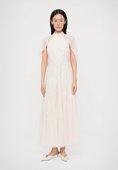 Biele tylové šaty s vysokým golierom a plášťovými rukávmi, zdobené textúrovanými ozdobami, ktoré plynú až do maxi dĺžky. Špicaté béžové opätky.