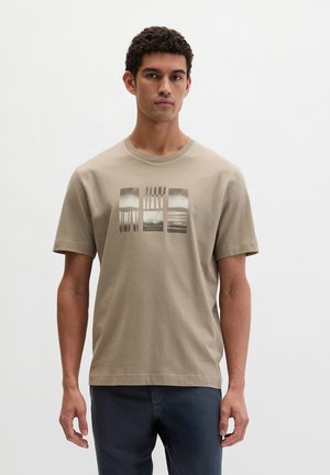 Mężczyzna w beżowej koszulce z nadrukiem i ciemnych spodniach, stojący na tle jednolitej białej ściany, zwrócony przodem z neutralnym wyrazem twarzy.