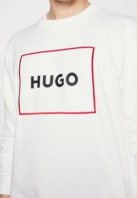 Sudadera blanca con un logo rectangular negro que dice "HUGO," bordeado con costura roja. Hecha de una tela suave y con cuello redondo.