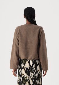 Osoba s pletenými vlasy, oblečená v hnědém texturovaném dlouhém rukávu a sukni s abstraktním vzorem v černé, bílé a zelené barvě, stojící zády.