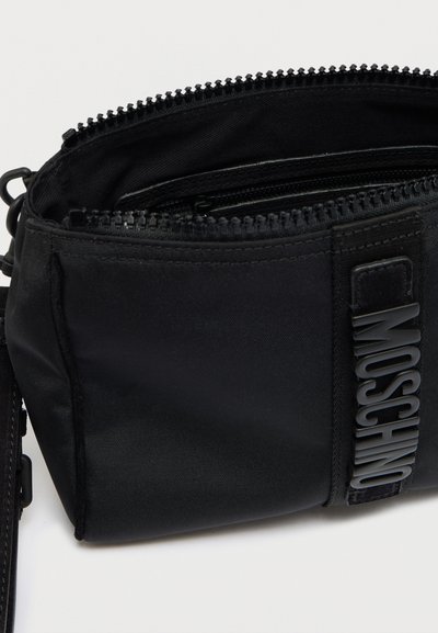 Μαύρο νάιλον τσάντα ώμου με φερμουαρ στα διαμερίσματα, ανάγλυφο λογότυπο και ρυθμιζόμενο ιμάντα. Συμπαγές ορθογώνιο σχήμα.