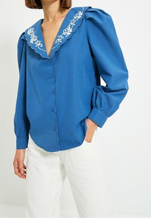 Blouse bleue avec des manches ballon, un col en dentelle ornée de broderies florales blanches, et une fermeture à boutons sur le devant. Portée avec un pantalon blanc.