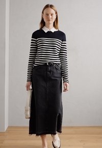 Csíkos pulóver fekete és fehér vízszintes csíkokkal, fekete farmer szoknya és világos színű loaferek kíséretében. Egyszerű fal háttér.