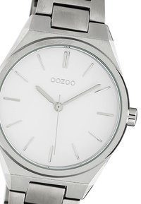 Montre en acier inoxydable avec un boîtier rond, un cadran blanc, des index horaires argentés et un bracelet métallique. Présente un design minimaliste et une texture lisse.