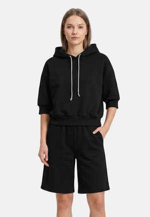 Jonge vrouw met een zwarte korte mouwen hoodie met trekkoorden en bijpassende zwarte shorts tot de knie, staand met één hand in de zak.