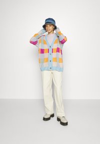 Cardigan tricoté coloré avec une base bleue, présentant de grands motifs à carreaux orange, rose et violet ; fermeture à boutons. Pantalons et chaussures de couleur crème.