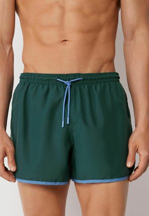 Man draagt donkergroene zwemshorts met lichtblauw trekkoordje en rand, staand tegen een effen lichte achtergrond.