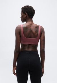 Γυναίκα με κοντά σγουρά μαλλιά, φορώντας ροζ αθλητικό σουτιέν και μαύρο κολάν, στέκεται με την πλάτη της προς τα εμπρός, μπροστά σε ένα απλό φόντο.