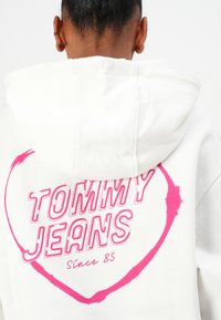 Haut à capuche blanc avec un graphique en cœur rose et le texte "TOMMY JEANS" avec "Depuis 85" en dessous. Tissu lisse, design décontracté, capuche visible.