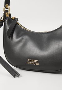 Black leather shoulder bag with gold-tone hardware, zipper closure, and embossed "TOMMY HILFIGER" logo accent. Smooth texture, curved shape.