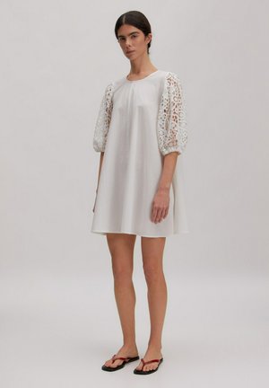 Vrouw staand in een korte witte jurk met kanten pofmouwen en rode teenslippers tegen een eenvoudige lichte achtergrond.