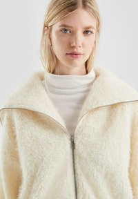 Giacca a zip color crema con una superficie morbida e testurizzata e un ampio colletto, indossata sopra un dolcevita bianco. Dettaglio della zip in argento visibile.