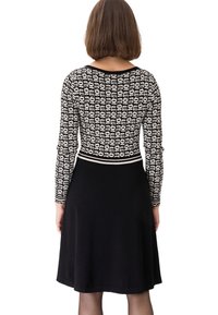 Femme aux cheveux courts et bruns portant un haut à manches longues à motif noir et blanc ainsi qu’une jupe noire unie, tournée dos au spectateur.