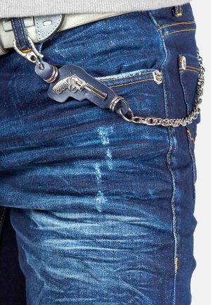 Blauwe denim jeans met een gestructureerde afwerking hebben een decoratieve metalen ketting en een nep-pistool sleutelhanger die aan de riemlus is bevestigd.