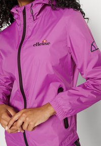 Person klädd i en ljuslila ellesse-jacka med svart dragkedja och logotyp, händerna knäppta vid midjan, lockigt hår synligt vid kragen.