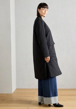 Abrigo ligero negro con mangas largas, bolsillos laterales y un ajuste holgado, combinado con pantalones de mezclilla de pierna ancha con un dobladillo deshilachado.