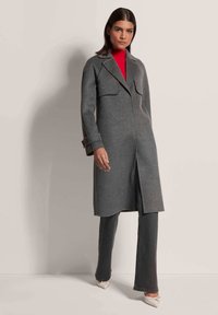 Grau melierter Wollmantel mit großen Revers und Fronttaschen, getragen über einem roten Rollkragenpullover und kombiniert mit dunklen Jeans und hellen Schuhen.