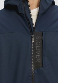 Giacca a vento blu navy con chiusura a zip, caratterizzata da una toppa di tessuto nera contrastante con il testo "S. OLIVER" e un cursore della zip con il marchio.