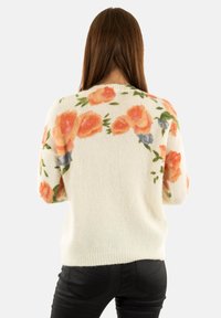 Mujer con cabello largo castaño que lleva un suéter blanco con patrones florales en naranja y verde, combinado con pantalones negros, de pie de espaldas frente al espectador.