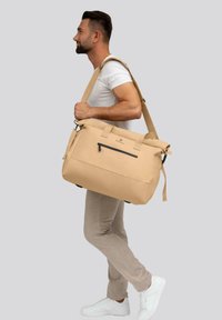 Borsa duffel beige realizzata in materiale resistente con una tasca con zip nera e tracolla regolabile. Forma rettangolare, dotata di manici superiori e design minimalista.