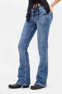 Žena v modrých bootcut džínoch, čiernych kožených čižmách a čiernom zberanom tričku s šnúrkami, stojí s jednou rukou v vrecku.