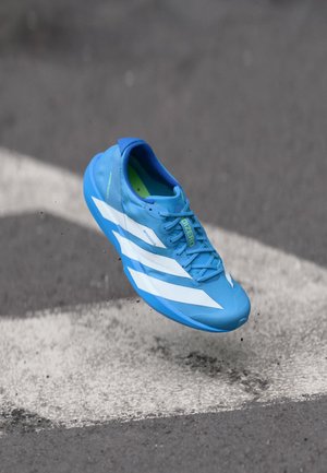 Blue running shoe with white stripes and "ADIZERO" text, captured mid-air above an asphalt surface with white road markings.