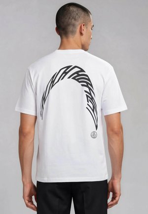 Mann mit kurzen Haaren steht mit dem Rücken zur Kamera, trägt ein weißes T-Shirt mit großem schwarzem, geschwungenem Grafikdesign und kleinem rundem Logo im unteren Rückenbereich.