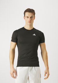 Camiseta atlética negra hecha de una tela suave y elástica; presenta mangas cortas y un logo blanco en el lado izquierdo del pecho. Diseño ajustado.