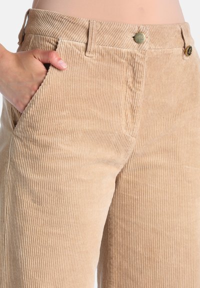 Main dans la poche d’un pantalon beige en velours côtelé taille haute avec bouton et passants de ceinture, montrant des rainures verticales texturées.