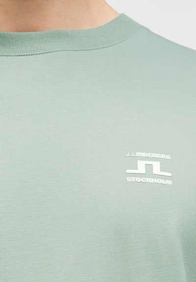 Κοντινό πλάνο στο λαιμόκοψη ενός ανοιχτού πράσινου πουκαμίσου με λευκό λογότυπο J. Lindeberg Stockholm στο στήθος.