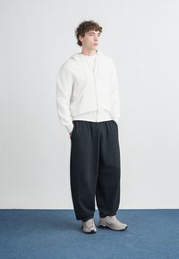 Λευκή ζακέτα με φερμουάρ, γκρι μπλουζάκι από μέσα, μαύροι άνετοι παντελόνια, και γκρι αθλητικά παπούτσια. Απλός σχεδιασμός χωρίς ορατά μοτίβα.