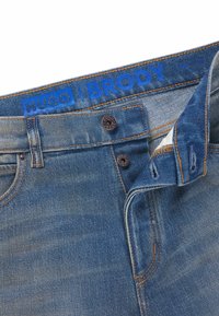 Denim jeans in medium blue with a faded texture, featuring copper hardware, orange stitching, and a blue logo waistband. Button and zip closure.