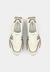 Lacoste tenisice u krem boji s sivim i crnim prugama, sa teksturiranim gumenim potplatom, okruglim vrhom i vezicama.